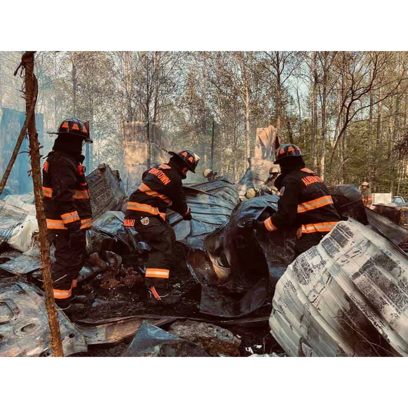 Firefighters in black Fire-Dex® turnouts with orange reflective striping searching through charred metal debris at a burned structure in a wooded area.