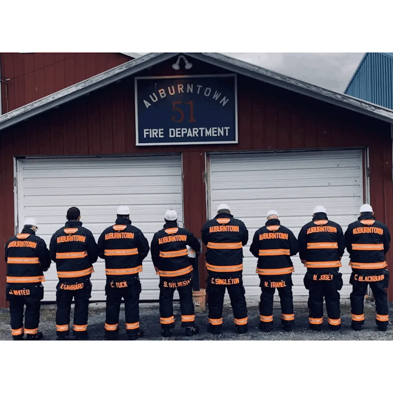 Eight firefighters in black Fire-Dex® turnouts with orange reflective striping standing with their backs to the camera in front of the Auburntown Fire Department station.