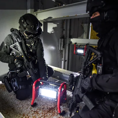 Two tactical officers in black helmets, face coverings, and gear holding rifles in a dim interior, lit by red Streamlight® Portable Scene Light II units with bright LED panels, one on the floor between them and another mounted on a ledge in the background.