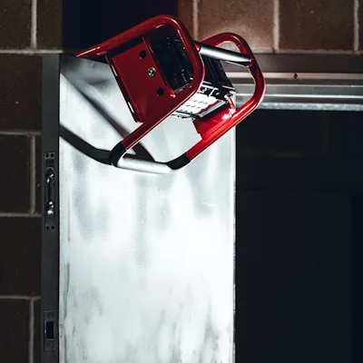 Red Streamlight® Portable Scene Light II hooked over the top edge of an open door beside a brick wall, its tubular roll-cage frame holding the LED panel angled downward to light a dark doorway.