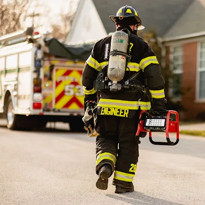 Firefighter in turnout gear and breathing apparatus, marked "Engineer" on the back, walking away down a residential street while carrying a red Streamlight® Portable Scene Light II by its handle, with a fire engine parked in the background.