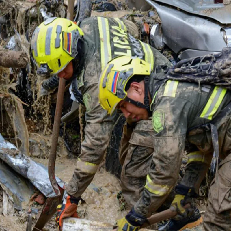 Team Wendy® EXFIL® SAR Backcountry™ helmet in use: two rescue workers in mud-covered uniforms and yellow-green safety helmets with flag patches digging through debris with shovels at a disaster site, with a wrecked vehicle and wreckage in the background.