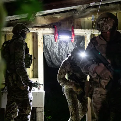 Three soldiers in camouflage uniforms and helmets, one with a mounted headlamp, entering a dark wooden structure at night, lit by a red Streamlight® Portable Scene Light II mounted overhead above the doorway.