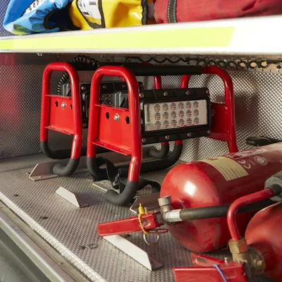 Red Streamlight® Portable Scene Light II units stored side by side in a fire apparatus compartment, their tubular roll-cage frames resting on a metal diamond-plate floor beside red fire extinguishers and gear bags on the shelf above.