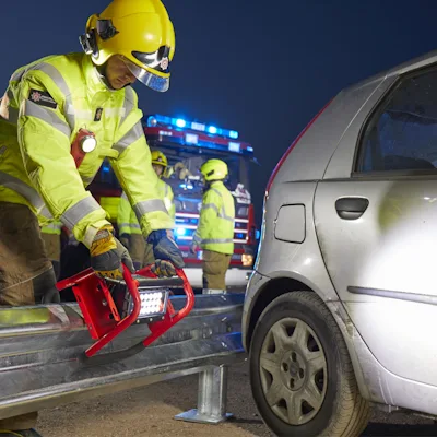 Firefighter in a yellow helmet and high-visibility gear setting a red Streamlight® Portable Scene Light II on a roadside guardrail beside a silver car at a night rescue scene, with a fire engine and crew in the background.