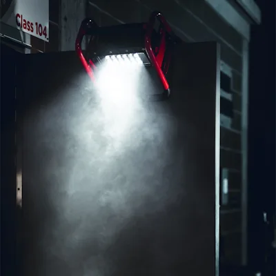 Red Streamlight® Portable Scene Light II hooked over the top edge of a door in the dark, its LED panel casting a bright downward beam through mist beside a "Class 104" sign.