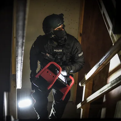 Police officer in a black helmet, face covering, and tactical gear carrying a red Streamlight® Portable Scene Light II up a dark stairwell, looking ahead while another light glows on the floor below.