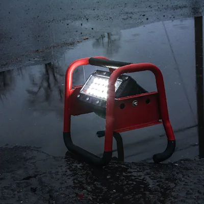 Red Streamlight® Portable Scene Light II with its 18-LED panel lit, set on wet pavement beside a large rain puddle at night, its tubular roll-cage frame and rubber-tipped feet holding it steady in the rain.