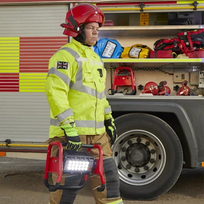 Firefighter in a red helmet and high-visibility yellow jacket carrying a red Streamlight® Portable Scene Light II with its LED panel lit, walking beside a fire engine with open equipment compartments and stored gear in the background.