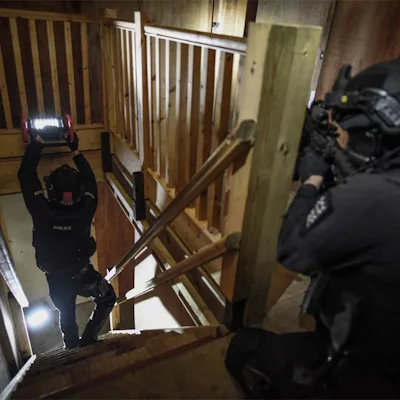 Police officer in a black helmet and tactical gear raising a red Streamlight® Portable Scene Light II with its LED panel lit while climbing a wooden stairwell, as a second armed officer in the foreground follows from behind.