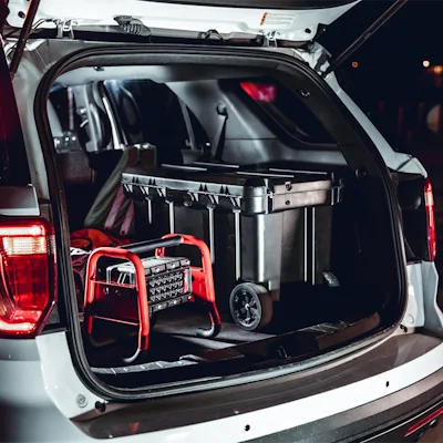 Red Streamlight® Portable Scene Light II stored in the open rear cargo area of a white SUV at night, resting beside a black wheeled equipment case with its tubular roll-cage frame and LED panel visible.