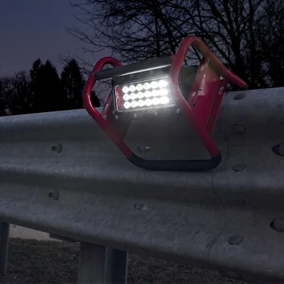 Red Streamlight® Portable Scene Light II with its 18-LED panel lit, hooked onto a metal highway guardrail at dusk with bare trees and a darkening sky in the background.