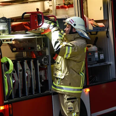 Firefighter in a white helmet and tan turnout gear with yellow reflective striping lifting a red Streamlight® Portable Scene Light II from an open fire truck equipment compartment filled with hoses and stored gear.