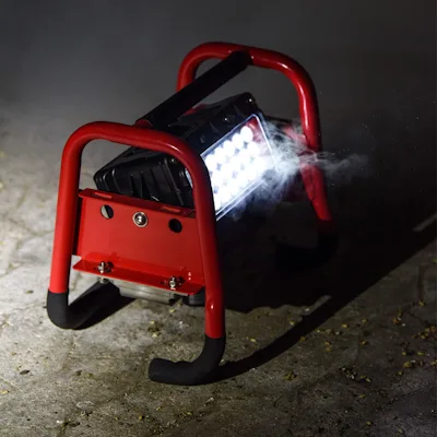 Red Streamlight® Portable Scene Light II with its 18-LED panel lit, sitting on dark pavement in its tubular roll-cage frame with rubber-tipped feet, casting bright light and wisps of mist in a dim outdoor setting.