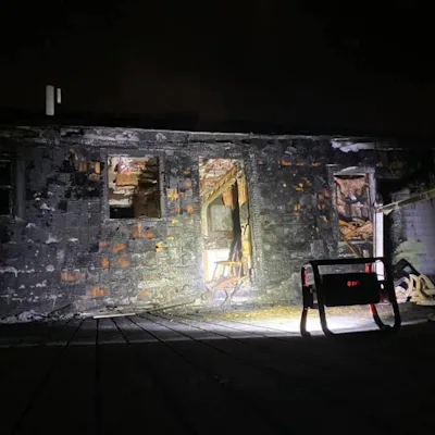 Red Streamlight® Portable Scene Light II facing a fire-damaged, soot-blackened brick building at night, its bright beam lighting the charred walls, broken windows, and open doorway.