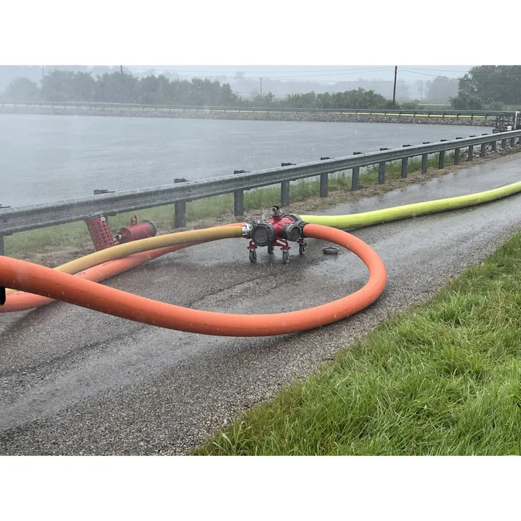 Orange, yellow, and lime-green BullDog™ Hi-Vol® firefighting hoses connected to a red manifold along a roadside guardrail beside a water source.