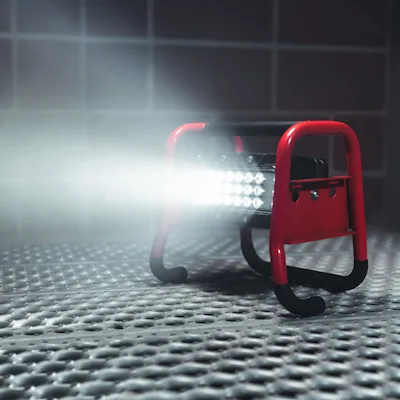 Red Streamlight® Portable Scene Light II in its tubular roll-cage frame sitting on a textured metal grate floor, with its 18-LED panel emitting a bright, wide beam across a dim tiled room.