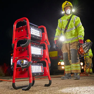 Firefighter in a yellow helmet, respirator mask, and high-visibility gear standing at a night rescue scene beside a stack of three red Streamlight® Portable Scene Light II units with their 18-LED panels lit, while a second firefighter operates a rescue tool in the background.