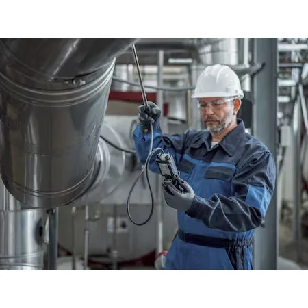 Worker in a hard hat, safety glasses, and blue coveralls using a Dräger X-am® 2800 Multi-Gas Detector with a sampling hose and probe near industrial ductwork.