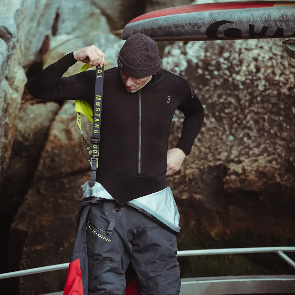 Man outdoors wearing Mustang Survival® Men’s Kazan Dry Suit Liner as thermal undergarments while putting on a dry suit over it, showcasing layered protection for cold water environments.