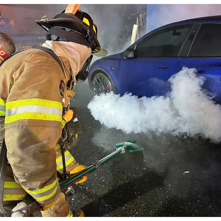 Firefighter using a Turtle Fire Systems™ EV Nozzle under a blue electric vehicle to release a dense cloud of water mist.
