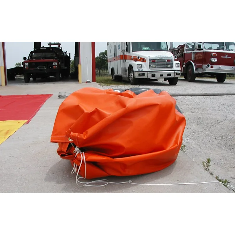 Folded orange Husky® self-supporting frameless portable water tank cinched shut with a drawstring on a concrete apron in front of fire station vehicles.