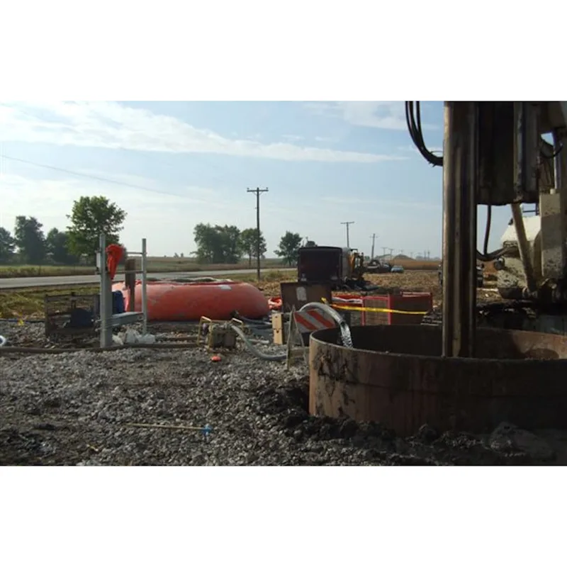 Orange Husky® self-supporting frameless portable water tank at a drilling site with a drill rig casing, hoses, and gravel ground in the foreground.