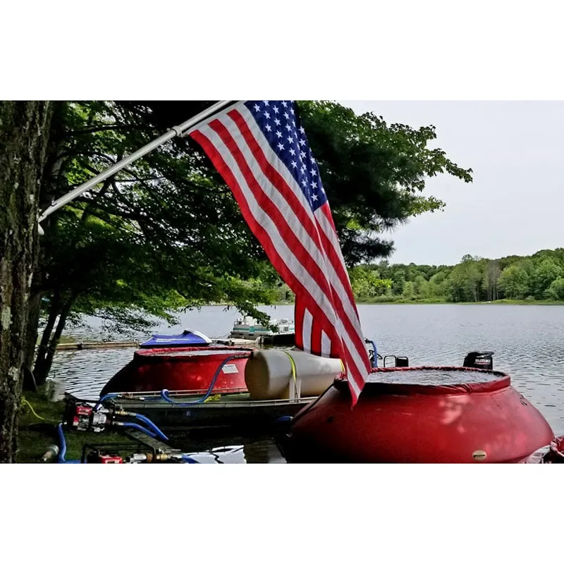 Red Husky® self-supporting frameless portable water tanks set up beside a lake with a pump, blue hoses, and an American flag in the foreground.