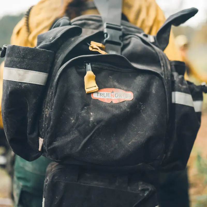 Front view of the black True North Gear Fireball™ Pack worn and carried by a firefighter, featuring adjustable straps and durable materials designed for comfortable, hands-free transport during emergency operations.
