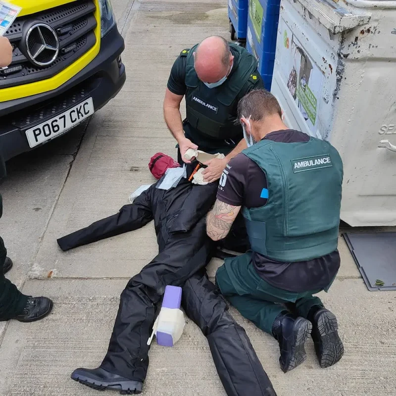 A Ruth Lee™ Duty Training Manikin lies on the ground while two EMS personnel provide medical attention, simulating a realistic emergency response scenario.