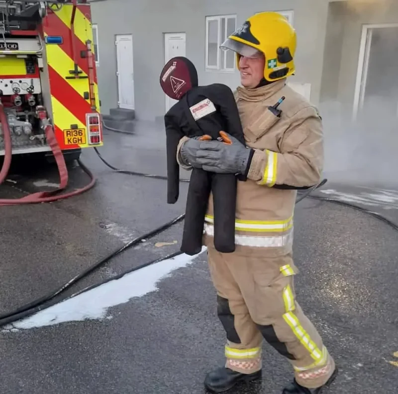 A toddler-size Ruth Lee™ Training Manikin is being carried by a firefighter in full gear, simulating the rescue of a small child during emergency response training.