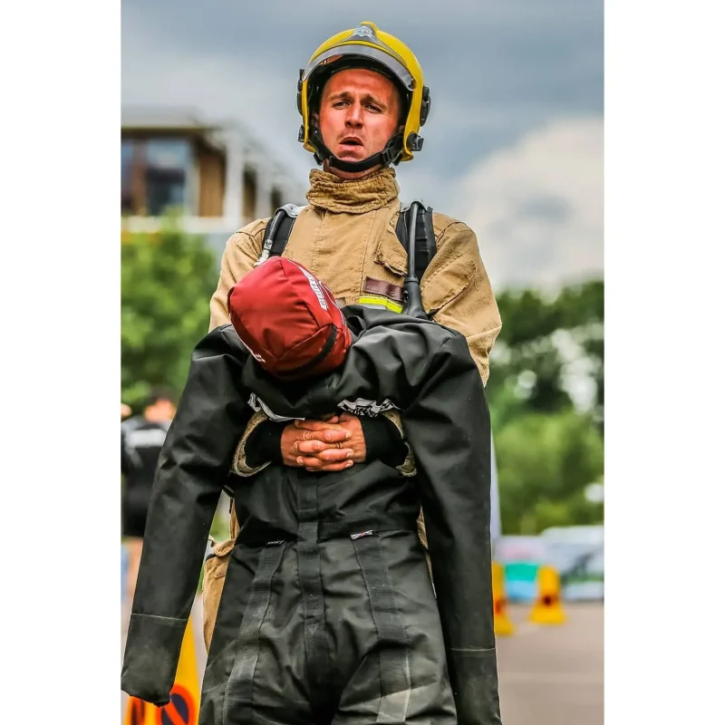 A close-up view shows a firefighter in full gear carrying a Ruth Lee™ Duty Training Manikin, simulating a realistic rescue during emergency response training.