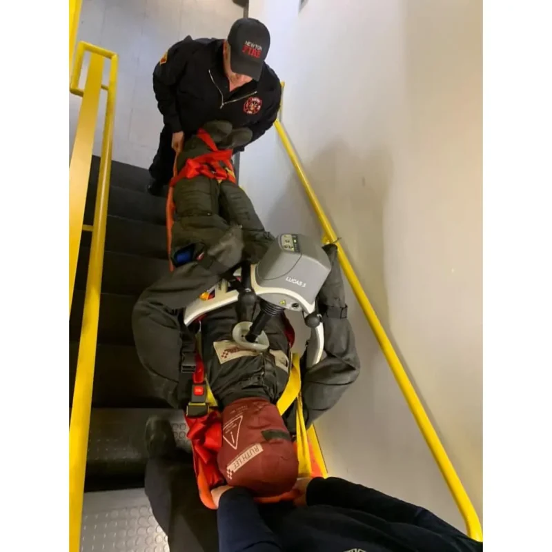 A Ruth Lee™ Duty Training Manikin is being transported down a staircase on a stretcher by firefighters during an emergency rescue training exercise.