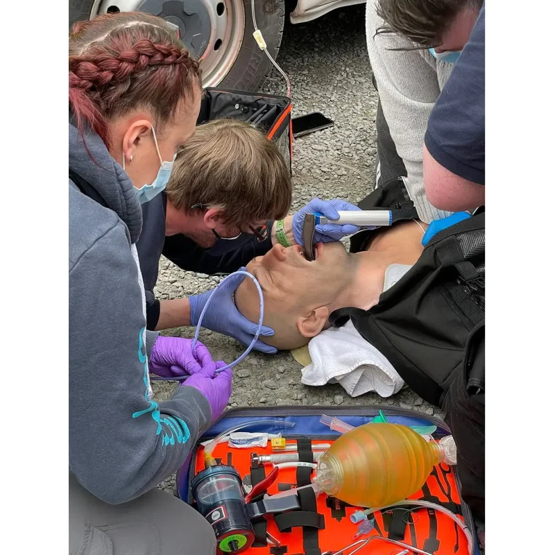 A Ruth Lee™ Advanced Water Rescue Manikin lying on the ground receiving CPR chest compressions and medical attention to the mouth during a simulated emergency rescue training.