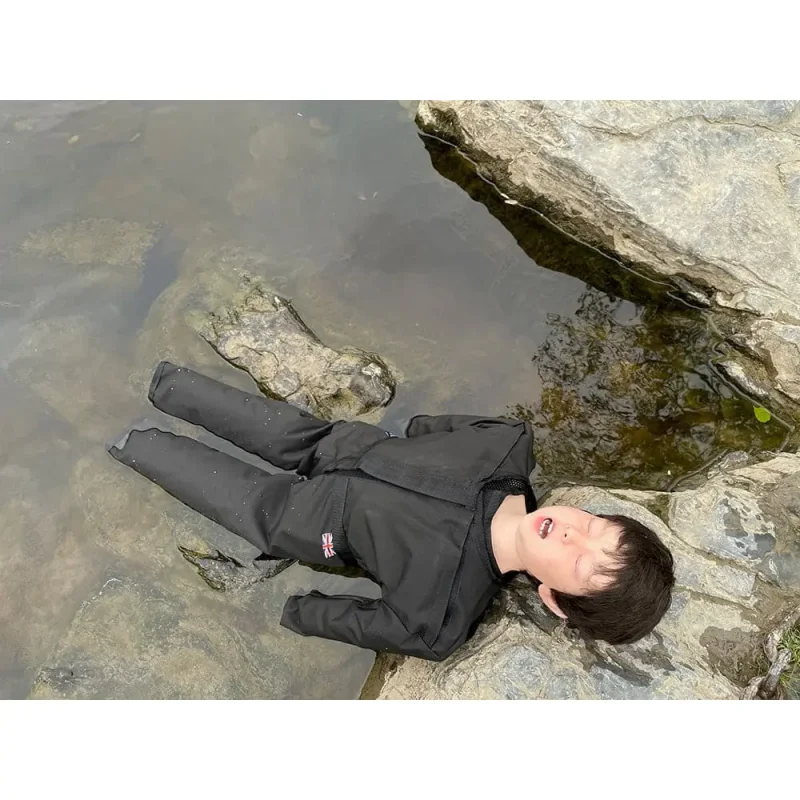 A Ruth Lee™ Advanced Water Rescue Toddler manikin lying on the rockside of a body of water, used for realistic child rescue training in aquatic environments.